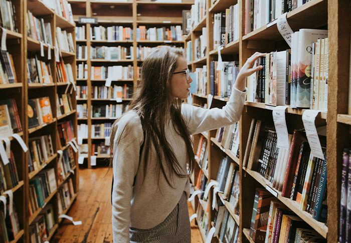 Woman with glasses browsing books in a library, focusing on learning skills to retire rich and improve life.