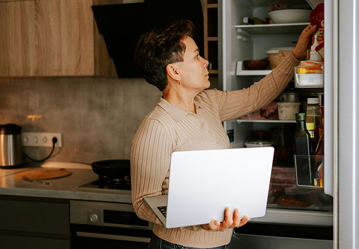 Woman holding a laptop and reaching into a refrigerator, representing easy skills to improve your life and retire rich.