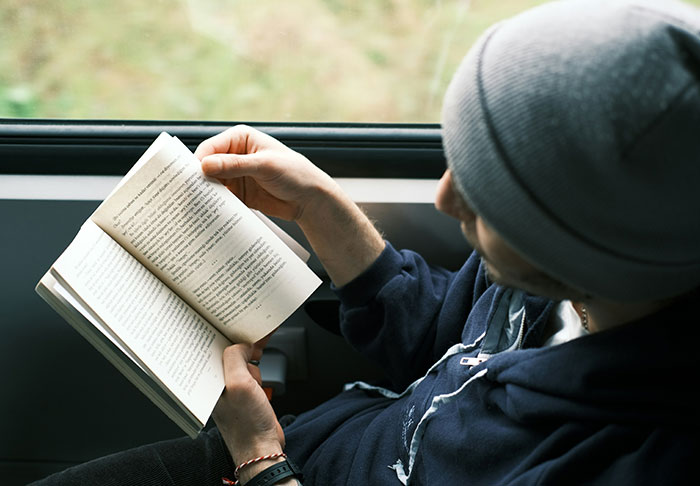 A young person wearing a beanie reading a book near a window, focusing on learning skills to improve life.