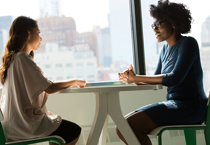Two women discussing easy skills to improve life while sitting at a table with a city view in the background.