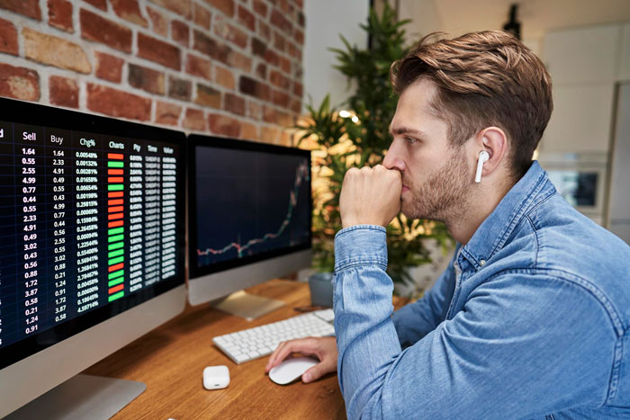 Unemployed guy sitting at desk, staring at dual monitors with stock charts, wearing AirPods, looking pensive