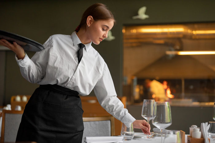 Unemployed Guy theme: young waitress in white shirt setting a restaurant table, placing a wine glass