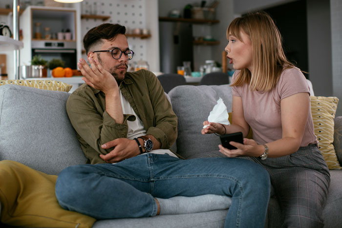 Unemployed guy on couch acting dismissive as friend holds phone and tissue, arguing in a living room Unemployed guy on couch acting dismissive as friend holds phone and tissue, arguing in a living room