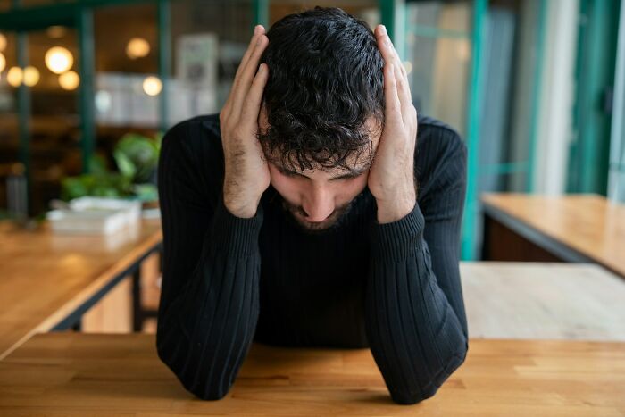 Man in a black sweater holding his head in panic at a wooden table, overwhelmed by accidental texts and messages.