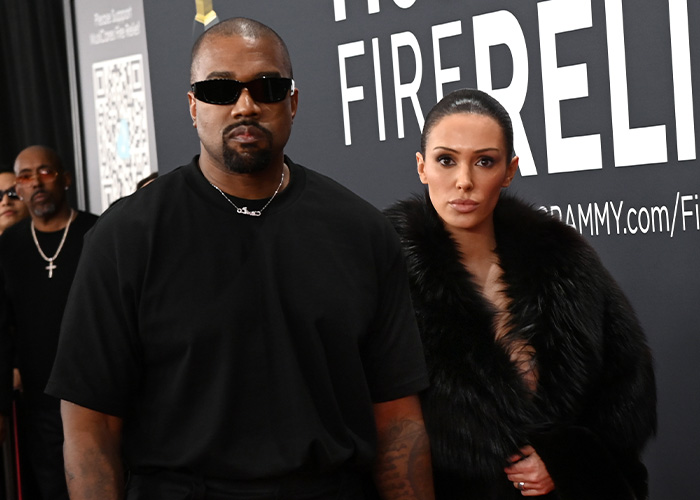 Bianca Censori wearing stylish pants and a black fur coat, standing next to a man in sunglasses at an event.