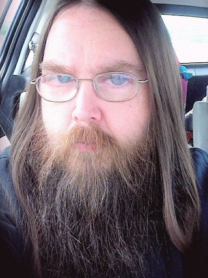 Man with long hair and beard wearing glasses inside a car, illustrating bilingual people catching gossip awkwardly.