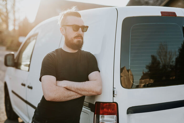 Bearded man wearing sunglasses leaning against white van, representing awkward bilingual gossip moments caught.