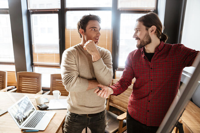 Two bilingual men in an office discussing and reacting to awkward gossip moments caught in conversation.