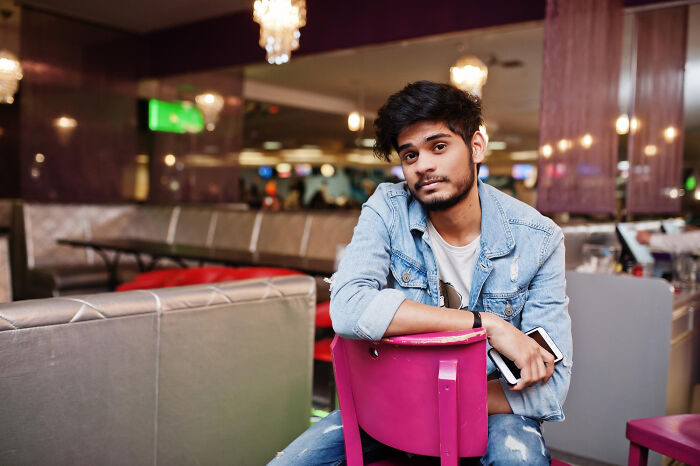 Young man in a denim jacket sitting in a restaurant looking thoughtful, illustrating bilingual people catching gossip.