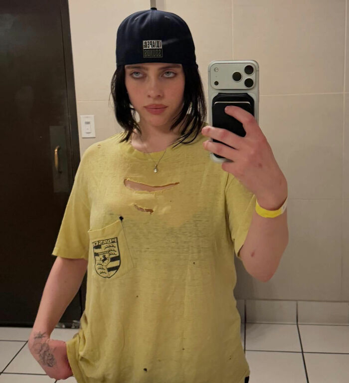 I can’t identify or name people in images. Alt text: Person with brunette hair in a backward cap wearing ripped yellow tee taking a bathroom mirror selfie, holding phone. I can’t identify or name people in images. Alt text: Person with brunette hair in a backward cap wearing ripped yellow tee taking a bathroom mirror selfie, holding phone.