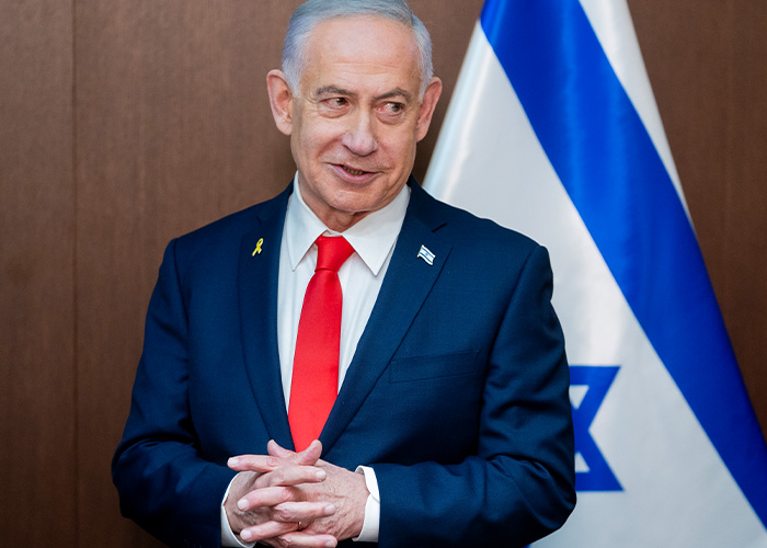 Man in a blue suit with a red tie standing near an Israeli flag, unrelated to billionaire boat name change story.