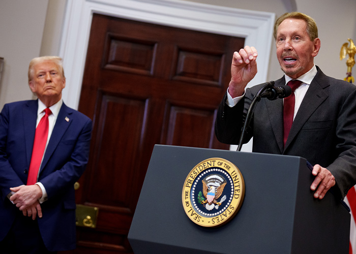 Billionaire speaking at podium with presidential seal while another man listens during formal event indoors