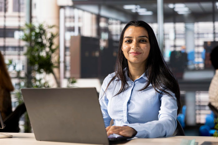 Young employee with South Asian ethnicity working on laptop in modern office, showing confident and hopeful expression.