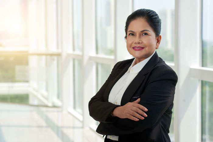Confident businesswoman in a black blazer standing with arms crossed by office windows, depicting bullied employee concept.