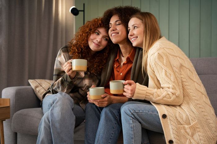 Three women friends cuddling on a sofa with mugs, smiling, illustrating boyfriend ban friends concerns Three women friends cuddling on a sofa with mugs, smiling, illustrating boyfriend ban friends concerns