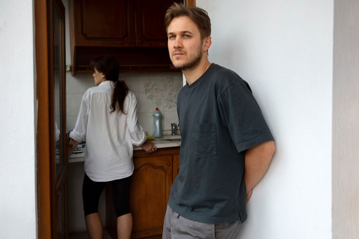 Man leaning in doorway looking upset while woman cooks, suggesting boyfriend ban friends conflict Man leaning in doorway looking upset while woman cooks, suggesting boyfriend ban friends conflict