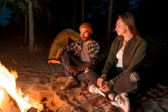 Couple sitting by a campfire at night outdoors, the man laughing while the woman looks on thoughtfully. Couple sitting by a campfire at night outdoors, the man laughing while the woman looks on thoughtfully.