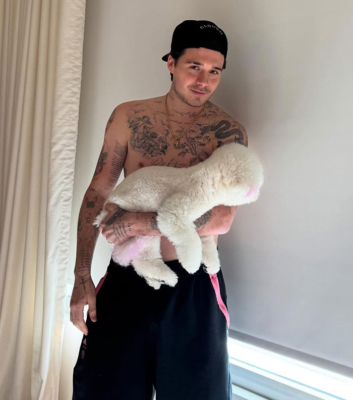 Brooklyn Beckham shirtless with tattoos, wearing a black cap, holding a fluffy white dog indoors by a window.