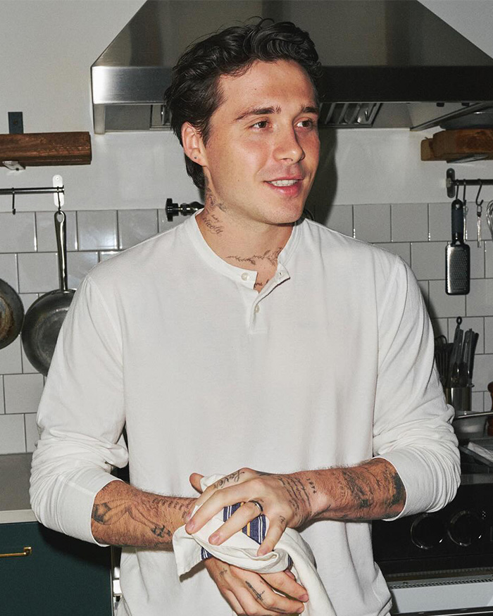 Brooklyn Beckham in a white shirt standing in a kitchen, showing tattoos on his neck and arms amid family drama.