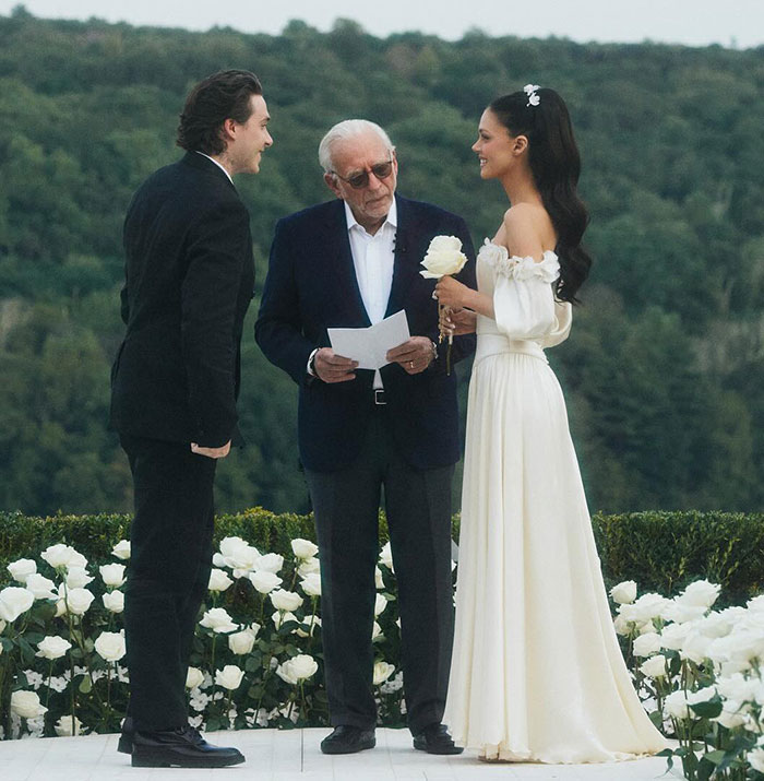 Brooklyn Beckham in a black suit standing with bride in white dress during outdoor wedding ceremony with white roses and forest background