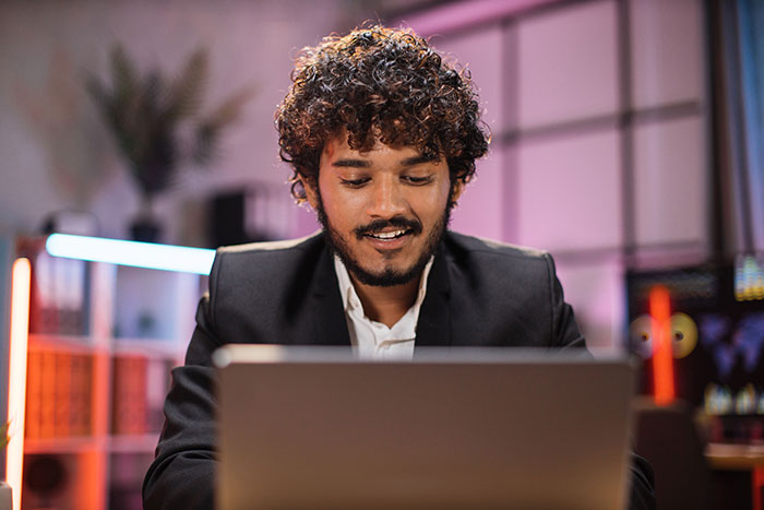 Young man with curly hair and beard working on a laptop in a dimly lit room, focused and smiling slightly.
