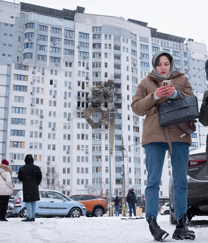 Woman in winter clothes using phone near damaged residential building in Ukraine amid tone deaf message backlash involving Timothée Chalamet fans. Woman in winter clothes using phone near damaged residential building in Ukraine amid tone deaf message backlash involving Timothée Chalamet fans.