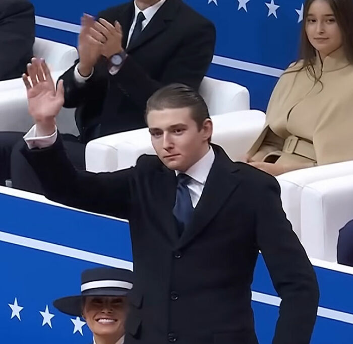 Young man in a suit waving while seated guests watch, related to Melania Trump and Mar-a-Lago rules controversy. Young man in a suit waving while seated guests watch, related to Melania Trump and Mar-a-Lago rules controversy.