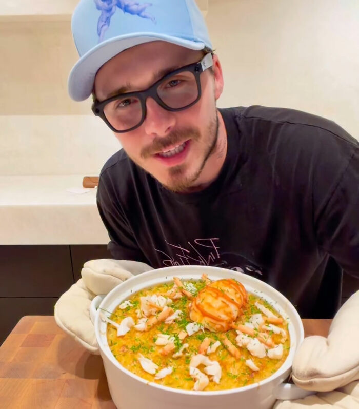 Brooklyn Beckham wearing glasses and a cap, holding a homemade breakfast dish in a kitchen setting. Brooklyn Beckham wearing glasses and a cap, holding a homemade breakfast dish in a kitchen setting.