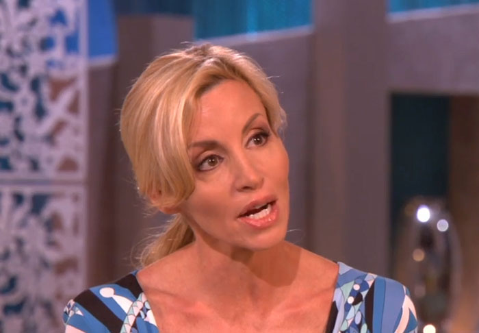 Blonde woman in a patterned blue dress speaking during a TV interview about celebrity infidelity scandals.