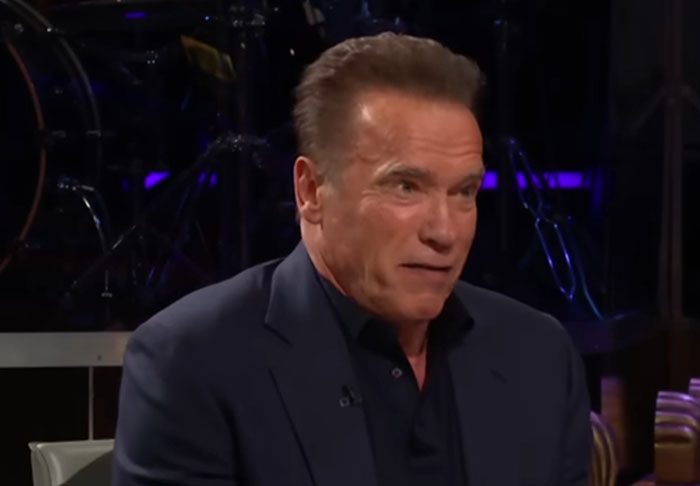 Actor in a dark suit discussing celebrity infidelity scandals during a televised interview in a studio setting.