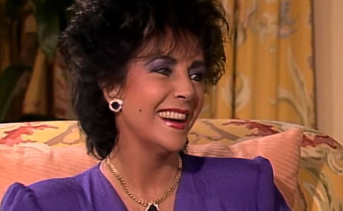 Woman in purple dress smiling during an interview about celebrity infidelity scandals on a patterned couch.
