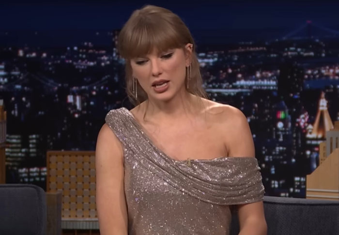 Celebrity wearing a glittery one-shoulder dress appearing out of touch with reality during a late-night talk show interview.