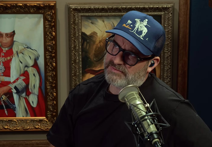 Man wearing a cap and glasses speaking into a microphone in a room with framed paintings behind him, celebrity out of touch.