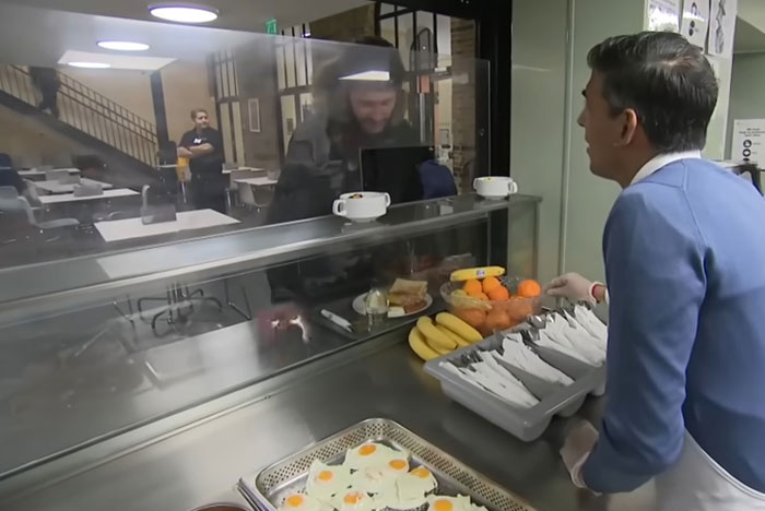 Celebrity out of touch with reality serving food behind a glass counter in a cafeteria setting with bananas and oranges visible.