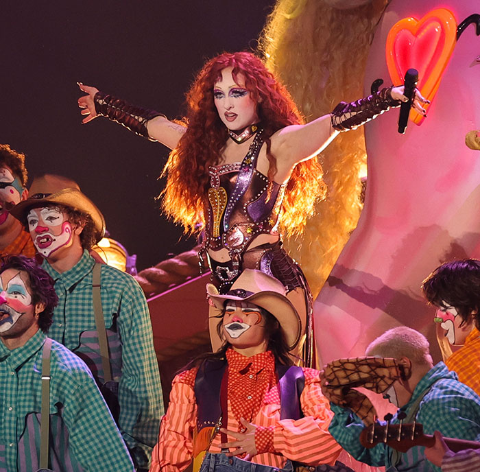 Performer in elaborate costume and makeup on stage surrounded by people in clown outfits during award show strict rules event.