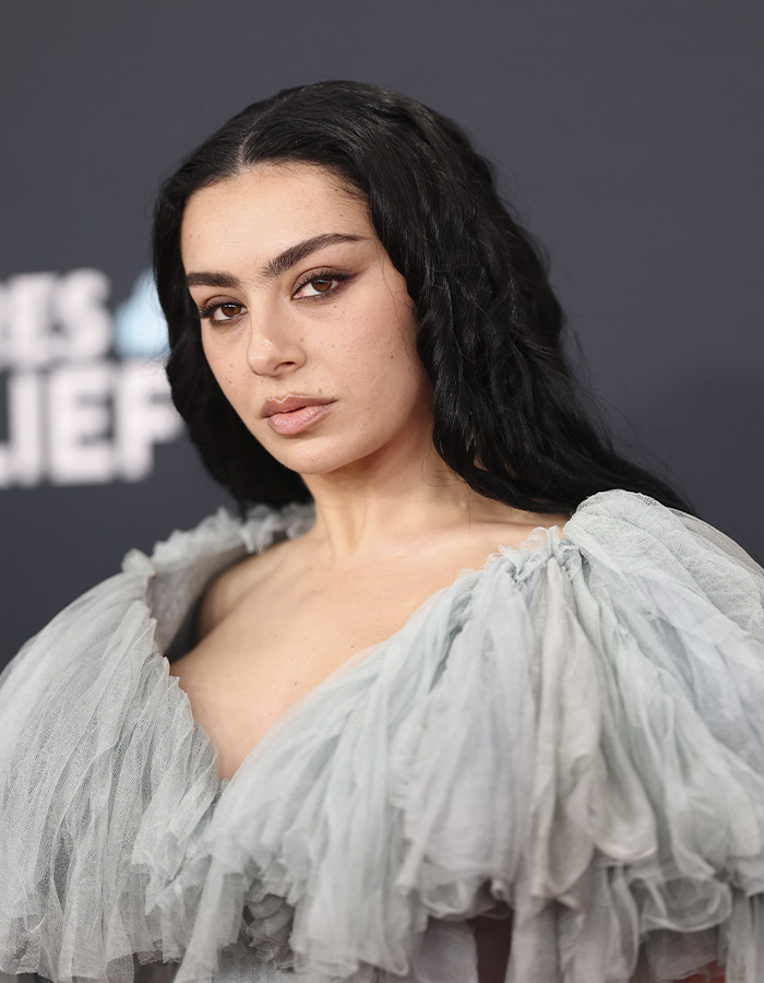 Charli XCX posing confidently in a racy braless photo dump with dark wavy hair and a sheer ruffled top.