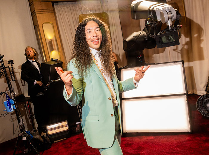 GlamBOT King wearing a pastel green suit, smiling and posing confidently at an event with lighting equipment behind him.