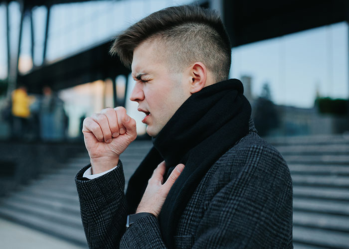 Young man outdoors coughing into his fist and clutching his chest, illustrating health signs of respiratory illness