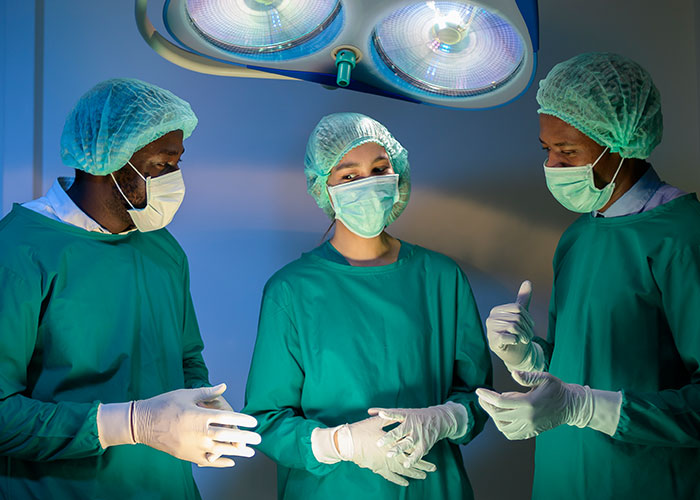 Three surgeons in operating room under bright surgical lights, wearing masks and gloves, discussing health signs