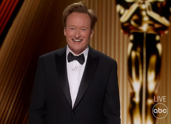 Male host in a tuxedo smiling on stage with a golden statue, illustrating strict rules celebrities follow during award shows.