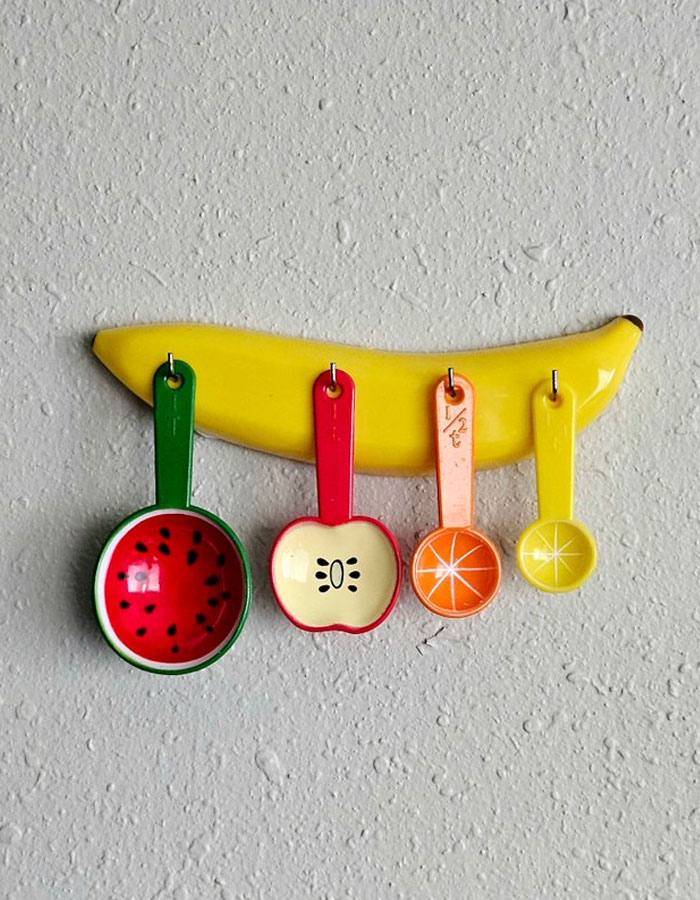 Colorful fruit-themed measuring spoons hanging on a banana-shaped wall hook as a cool interior design idea.
