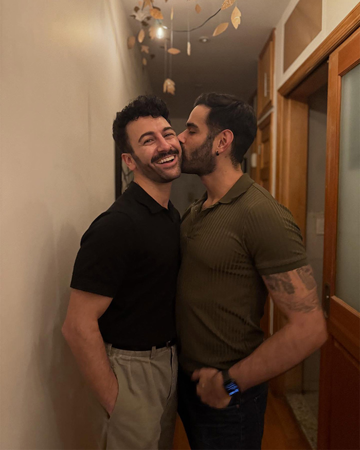 Gay couple in hallway, one kissing partner's cheek as they smile and embrace Gay couple in hallway, one kissing partner's cheek as they smile and embrace