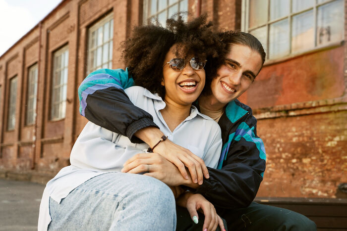 Two smiling teens embracing outside a brick school building, casual friends, high school secrets vibe