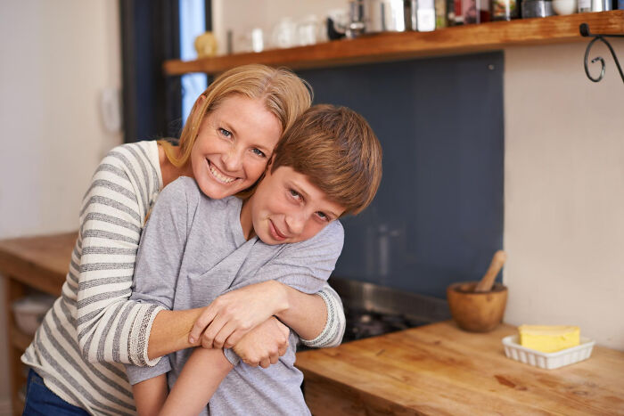 Smiling mother hugging teenage son in kitchen, warm family moment; high school secrets