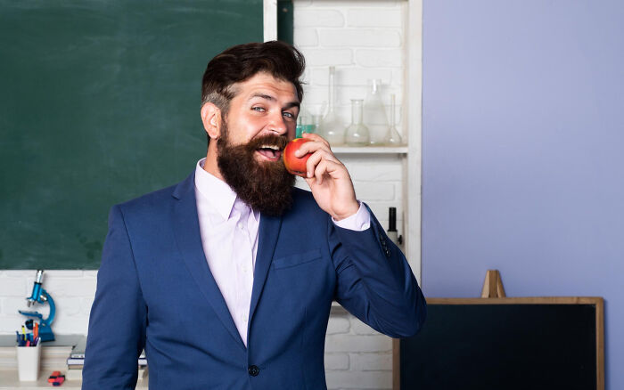 Bearded teacher in suit playfully biting an apple in classroom, hinting at high school secrets