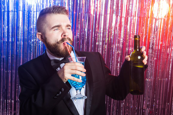 Bearded man in tuxedo at party sipping blue cocktail and holding wine bottle, high school secrets.