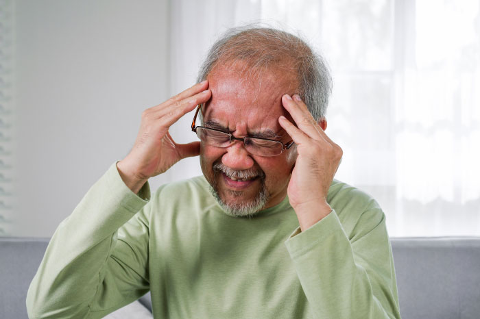 Elderly man in green shirt grimacing and holding temples, expressing stress related to MIL poisoning family dog incident.