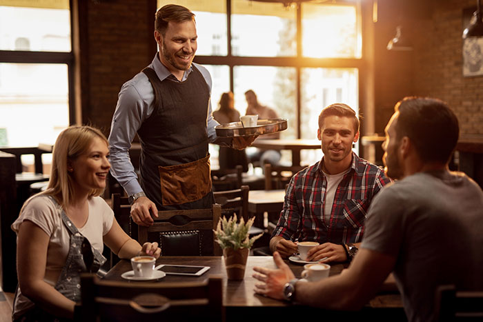 A male bestie at a caf&eacute; with his girlfriend and boyfriend, waiter serving coffee in a warm, sunlit setting.