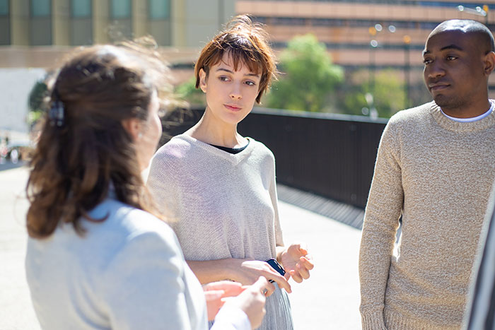 Three people having an outdoor conversation, displaying sharp intelligence and thoughtful expressions in a sunny setting.