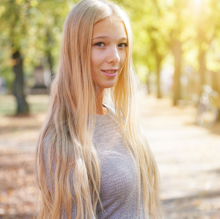 Young woman with long blonde hair smiling outdoors in sunlight, representing sharp intelligence that amazes and surprises.
